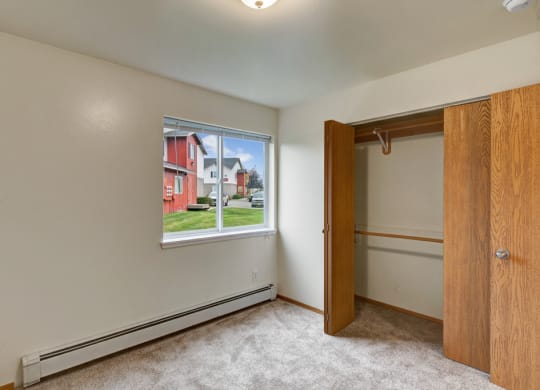Small, empty room here at Strawberry Lane Apartments with beige walls and carpet, open wooden closet doors, and a window showing green grass and red buildings outside, creating a calm tone.