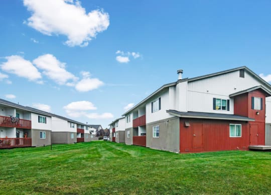 Stawberry Lane Apartment buildings with red and beige exteriors line a green grassy area under a clear blue sky with fluffy clouds, conveying a peaceful atmosphere.