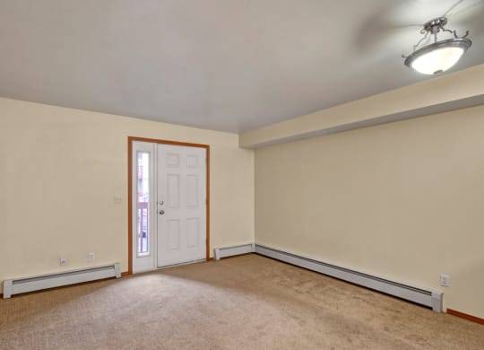 Empty living room here at Strawberry Lane Apartments with beige walls and carpet, featuring a white door with a side window. A round ceiling light casts a soft glow, creating a serene atmosphere.