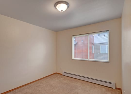 Small, empty beige room here at Strawberry Lane Apartments with carpeted floor, single window with blinds, and a ceiling light. The room has a simple, minimalist appearance.
