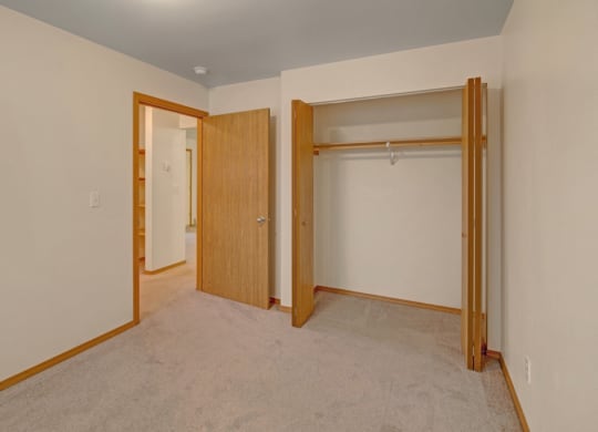 Empty beige room here at Strawberry Lane Apartments with an open closet on the right and a wooden door on the left. Soft carpet flooring, simple and clean atmosphere.