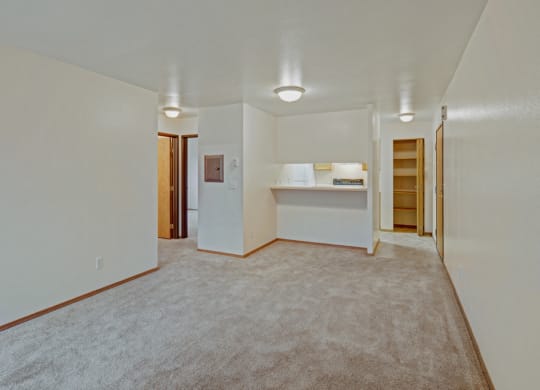 Spacious empty living room here at Strawberry Lane Apartments with light beige carpet. An open doorway leads to a lit, compact kitchen. The interior is bright and minimalistic.
