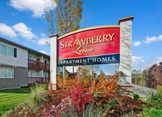 Sign for "Strawberry Lane Apartment Homes" amidst colorful shrubs, with an apartment building and a clear blue sky in the background. Bright, inviting scene.