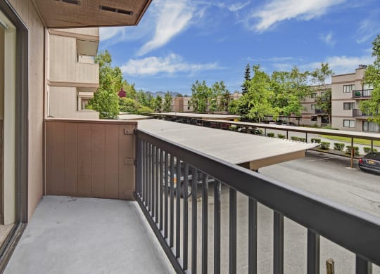 A small balcony here at Susitna Ridge Apartments with a metal railing overlooks a parking lot and covered parking. Surrounding are beige apartment buildings and green trees under a blue sky.
