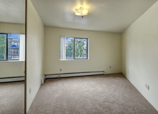Empty bedroom here at Susitna Ridge Apartments with beige carpet, cream walls, and a ceiling light. A window reveals green trees outside. The space looks clean and inviting.