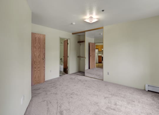 Empty room here at Susitna Ridge Apartments with beige carpet, mirrored closet doors reflecting a kitchen, wooden doors leading to a bathroom; minimalistic and bright atmosphere.