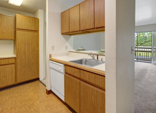 Compact kitchen here at Susitna Ridge Apartments with brown wooden cabinets, white countertops, and a sink. A glimpse of a carpeted living area with sliding glass doors to a balcony. Cozy atmosphere.