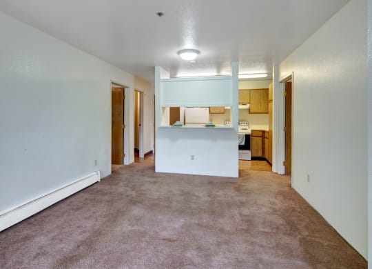 Spacious, empty apartment living room here at Susitna Ridge Apartments with beige carpet and white walls, a hallway with wooden doors, and a small kitchen with light wood cabinets.
