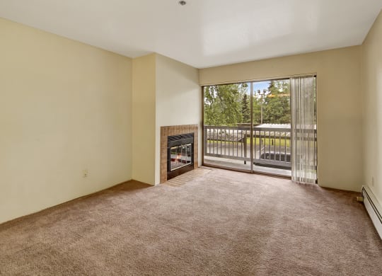 Empty living room here at Susitna Ridge Apartments with beige carpet and walls, featuring a tiled fireplace. A large sliding glass door leads to a balcony with views of greenery. Bright and serene atmosphere.