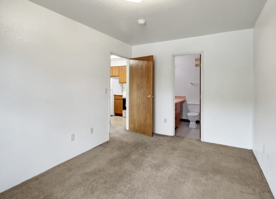 Empty bedroom here at Susitna Ridge Apartments with beige carpet and white walls, featuring open wooden doors leading to a kitchen with wooden cabinets and a bathroom. Bright, neutral tone.