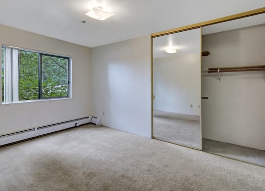 Empty, bright bedroom here at Susitna Ridge Apartments with beige carpet, large window showing greenery, and mirrored sliding closet doors reflecting ceiling light, creating a serene atmosphere.