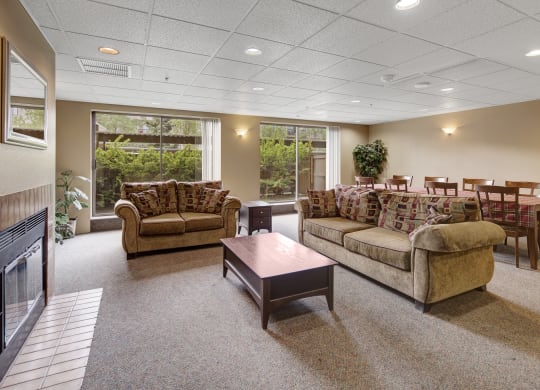 Spacious resident lounge here at Susitna Ridge Apartments with two beige sofas, a wooden coffee table, and a fireplace. Large windows reveal lush greenery. Dining area with a table, chairs, and a potted plant. Cozy and inviting tone.