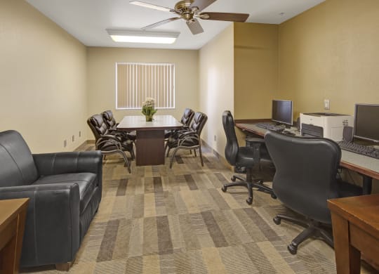 Susitna Ridge Apartment's modern, neutral-toned office features a round table with six leather chairs, a black sofa, two computers on desks, and a window with blinds.