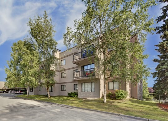 Three-story beige Sustitna Ridge Apartment building surrounded by lush green trees under a clear blue sky. Balconies feature potted plants, creating a serene atmosphere.