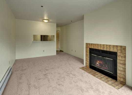 Empty living room here at Susitna Ridge Apartments with beige carpet, featuring a lit fireplace with a brown tile surround on the right, and a small serving hatch on the left wall. Cozy ambiance.