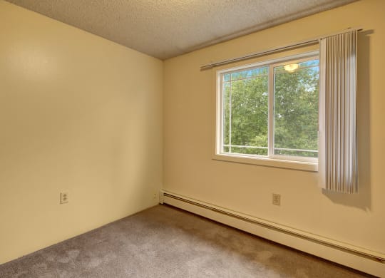 Empty bedroom here at Taiga Apartments with beige walls, carpeted floor, and a large window with vertical blinds. The window reveals lush green trees outside, conveying tranquility.
