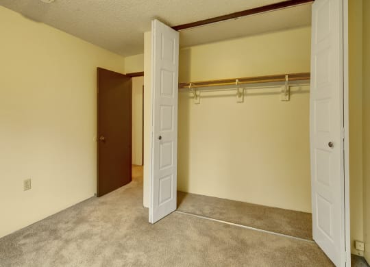 Empty beige room here at Taiga Apartments with open closet doors, revealing a wooden rod and shelf inside. Carpeted floor and an open brown door leading to another room. Quiet and clean atmosphere.