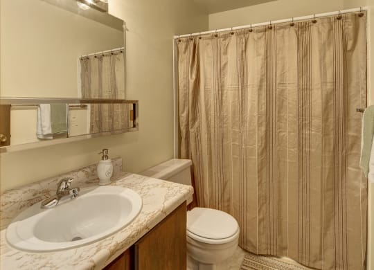 A small bathroom here at Taiga Apartments with a marble-patterned sink, soap dispenser, and toothbrush holder. Beige shower curtain, wooden door with towels, bright lighting.