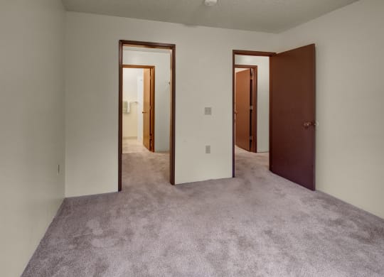 Empty bedroom here at Taiga Apartments with beige carpet, white walls, and open brown doors leading to two additional rooms. The atmosphere is neutral and minimalistic.