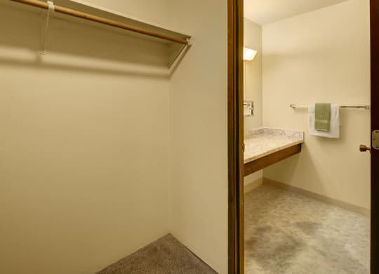 Minimalist interior here at Taiga Apartments showing an empty closet with a wooden rod and shelf. Adjacent bathroom features a marble countertop and towel rack with towels.