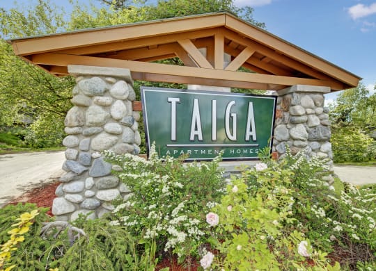 Stone sign for Taiga Apartment Homes amid lush green foliage and blooming flowers, set against a bright blue sky, conveying a welcoming and serene atmosphere.