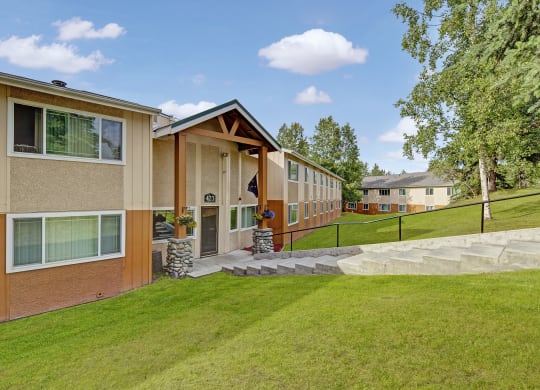 Two-story Taiga Apartment building with beige and orange siding, large windows, and a path leading up to the entrance. Lush green lawn and trees surround it.