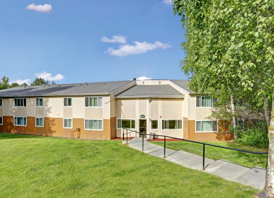 Two-story beige Taiga Apartment building with green-trimmed windows under a clear blue sky. A tree and lush grass flank the entrance, creating a serene atmosphere.