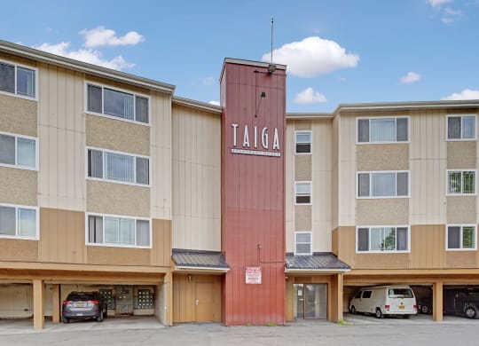 Four-story Taiga Apartment building with a vertical red sign reading "TAIGA." Beige facade with multiple windows and cars parked in ground floor garages.