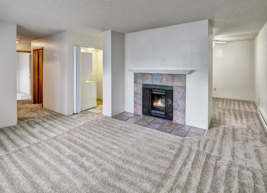 A spacious living room here at Terrace on the Lake Apartments with beige carpet features a lit fireplace with a tiled mantel. The room is bright, with white walls and a cozy feel.