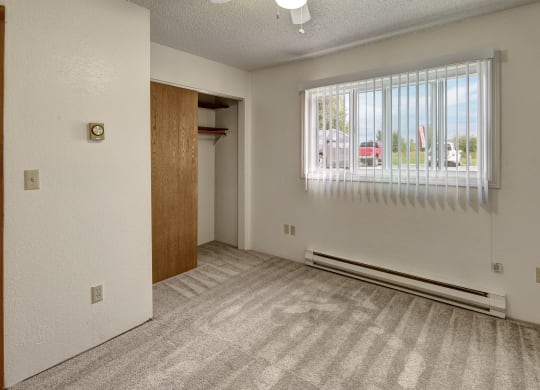 Empty bedroom Terrace on the Lake Apartments with beige carpet, white walls, and vertical blinds on a window. An open closet with a wooden door is on the left, creating a neutral, clean atmosphere.