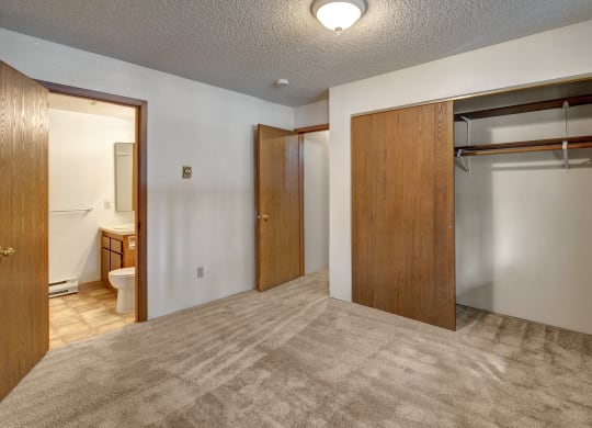 Empty bedroom here at Terrace on the Lake Apartments with beige carpet, a wooden closet with sliding doors, and an open door revealing a bathroom. Soft lighting creates a neutral, calm tone.