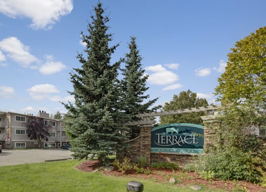 Apartment complex entrance with stone sign reading "Terrace on the Lake," flanked by tall evergreen trees. A building and parked cars are visible under a bright blue sky.