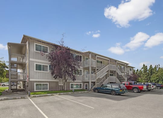 Three-story beige Terrace on the Lake Apartment building with external staircases, parked cars in front, and a cloudy blue sky. Trees frame the scene, offering a serene feel.