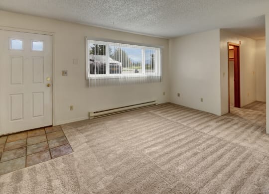 Empty living room here at Terrace on the Lake Apartments with beige carpet, white walls, and a light blue door with windows at the top. Large window with vertical blinds lets in natural light.