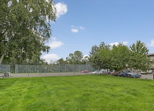 Lush green lawn in the foreground, fenced tennis court to the left, and parked cars to the right. Clear blue sky and leafy trees create a serene atmosphere here at Terrace on the Lake Apartments.