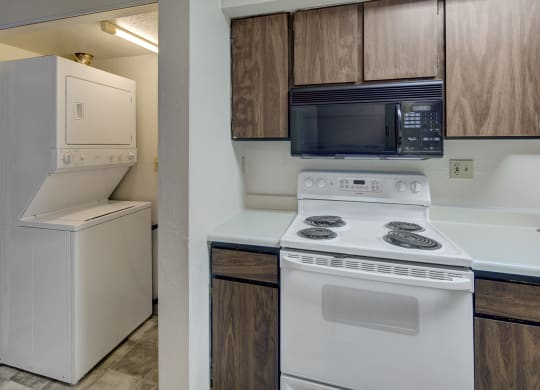 Compact kitchen here at Terrace on the Lake Apartments with white stove, dark wood cabinets, and built-in microwave. Adjacent, a stacked washer and dryer in a small laundry nook.