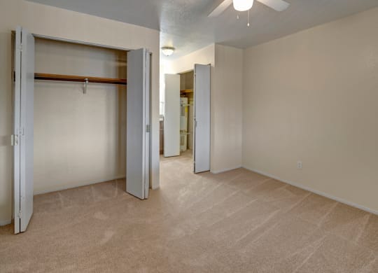 Empty beige bedroom here at The Club at Eagle Pointe Apartments with open closets revealing a wooden rod and shelves. Carpeted floor, a ceiling fan, and soft lighting create a neutral, spacious feel.