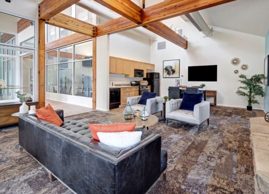 Resident Lounge here at The Highlands Luxury Apartments with high wooden beam ceilings, featuring a black leather sofa with colorful cushions, two white armchairs, and a stone fireplace. Walls have large windows, a wall-mounted TV, and art. The kitchen is in the background with light wood cabinetry. The room exudes warmth and sophistication.