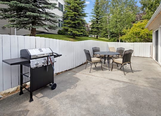 A patio here at The Highlands Luxury Apartments with a barbecue grill and a round table surrounded by four chairs. A white fence and trees surround the area, creating a peaceful setting.