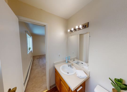 Bright bathroom here at the Village at Calais Apartments with beige walls, a wood cabinet sink, and a large mirror. A corridor with carpet and a window leads to another room.