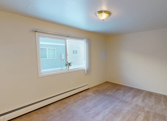 Spacious, empty bedroom here at the Village at Calais Apartments with beige carpet and walls, large window with vertical blinds, and a small plant on the sill. Overhead light fixture provides a cozy feel.