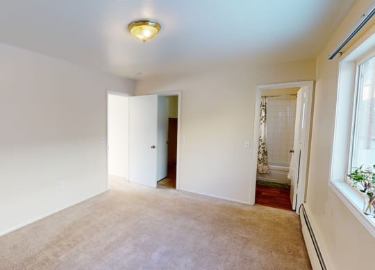 Empty bedroom here at the Village at Calais Apartments with beige carpet, cream walls, and a ceiling light. Open doors reveal a closet and a bathroom with a white tiled shower curtain. A large window adds brightness.