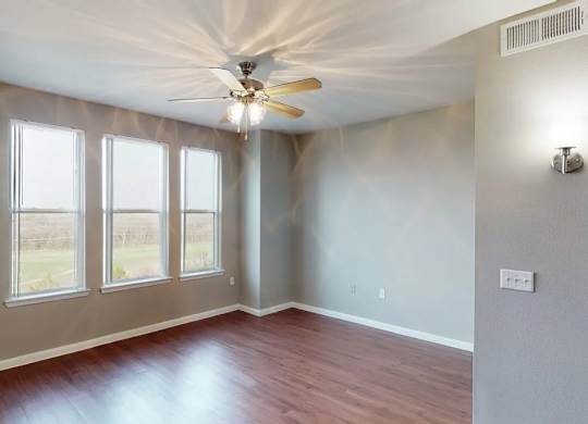 A bright empty living room here at BRIO at Firewheel with wood-style flooring, three tall windows with natural light, neutral walls, and a ceiling fan in an open, airy layout.