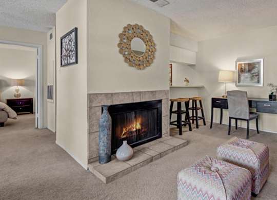 A cozy living room here at Cypress Pointe with soft beige carpet, neutral walls, and a lit fireplace creating a warm focal point. An open layout includes a built-in breakfast bar with seating and a small desk area, offering a comfortable and functional space for everyday living.