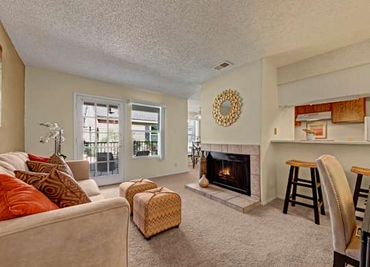 A warm, inviting living room here at Cypress Pointe with soft beige carpet, neutral walls, and a cozy fireplace serving as a central feature. An open layout connects to the kitchen with a breakfast bar and seating, while glass doors bring in natural light and provide access to a private patio.