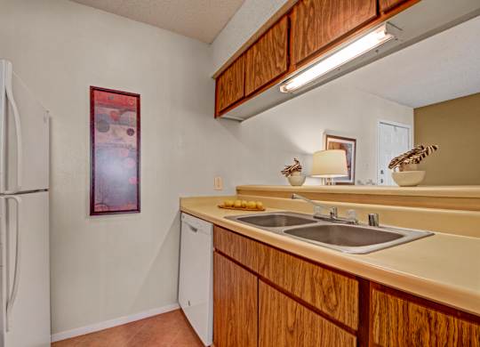 A compact kitchen here at Cypress Pointe with warm wood cabinets, beige countertops, and a double stainless steel sink beneath bright overhead lighting. White appliances, including a refrigerator and dishwasher, add a clean, functional feel to this cozy, practical cooking space.