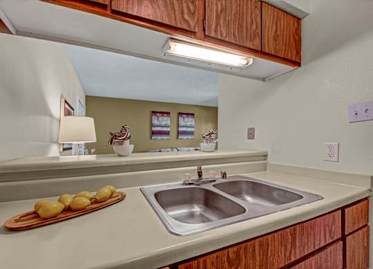 A functional kitchen here at Cypress Pointe with warm wood cabinets, beige countertops, and a double stainless steel sink beneath bright overhead lighting. An open pass-through overlooks the living area, creating a connected and practical layout for everyday use.