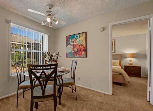 A bright dining area here at Cypress Pointe with beige carpet, neutral walls, and a large window with vertical blinds bringing in natural light. A ceiling fan adds comfort, while the open layout offers a clear view into the bedroom, creating a warm and inviting space for everyday dining.