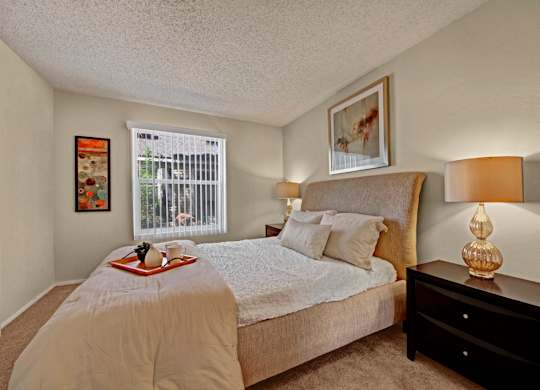 A comfortable bedroom here at Cypress Pointe with plush beige carpet, neutral walls, and a large window with vertical blinds allowing natural light to fill the room. A neatly staged bed with soft bedding and matching bedside lamps creates a calm, cozy, and inviting atmosphere.