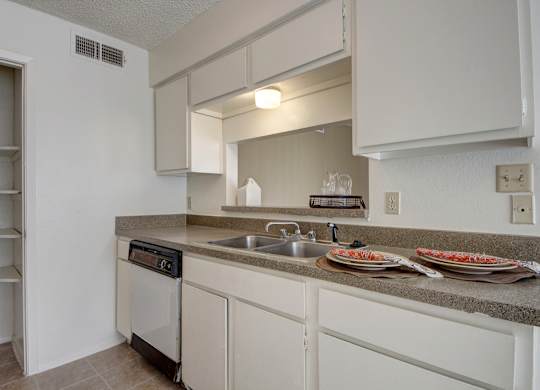 A clean kitchen here at Fairgreen with white cabinets, neutral countertops, and a double stainless steel sink creating a bright, practical workspace. A dishwasher and open pass-through to the living area add convenience and an easy, connected layout.